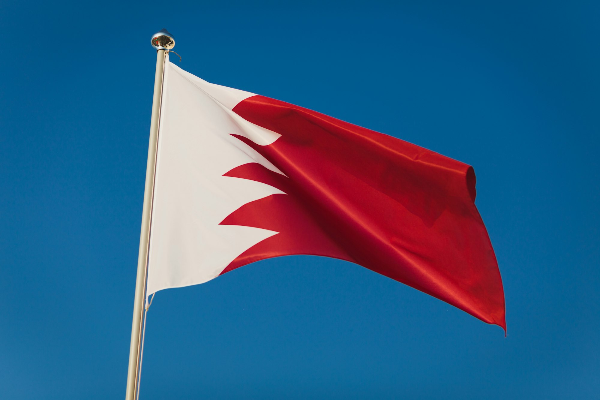 Red and white Bahrain flag, capital Bruxelles. National flag on flagpole in front of blue sky